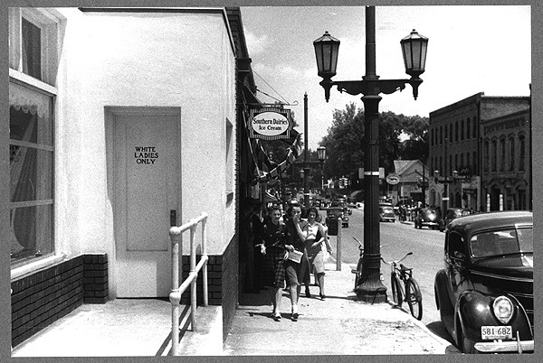 A bathroom sign reads "White ladies Only." The restroom sits beside a busy downtown road, and there are buildings, pedestrians, and cars in the setting. Black and white photo. 